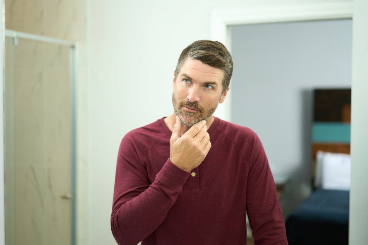 A man in a red shirt looking into a mirror and fixing his beard